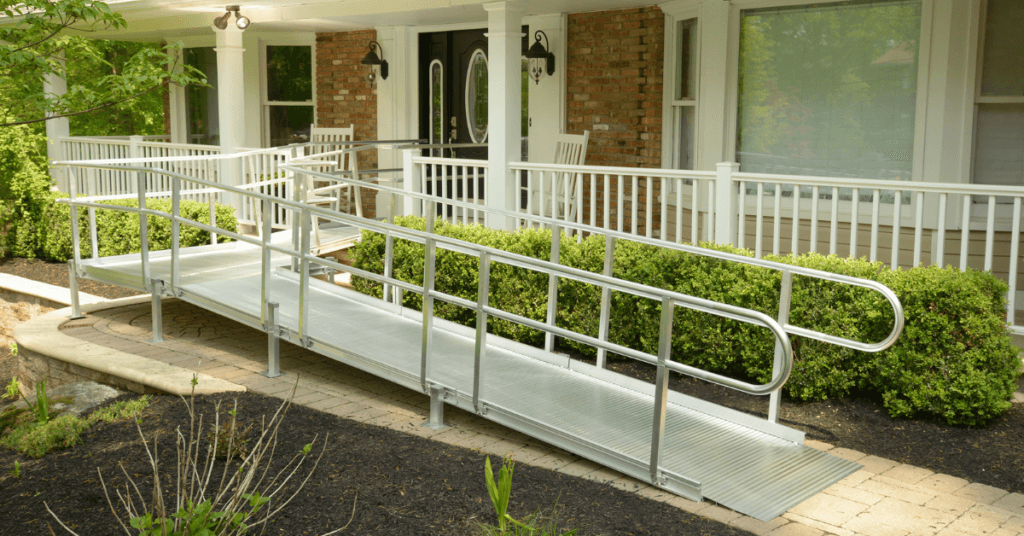 A metal wheelchair ramp leads to the front porch of a house, bordered by bushes and a brick wall.