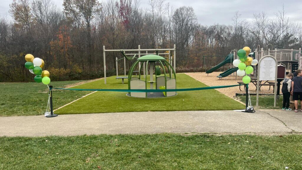 A playground with green and yellow balloons, featuring a climbing structure and slide, is cordoned off with a blue ribbon, with several people nearby.