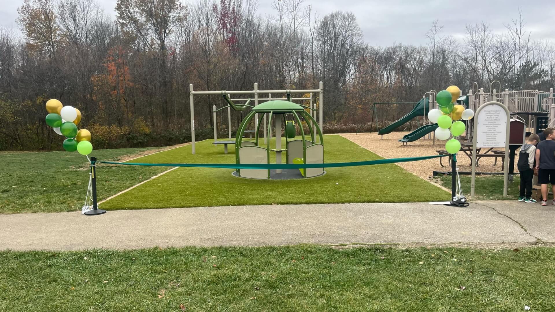 A playground with green and yellow balloons, featuring a climbing structure and slide, is cordoned off with a blue ribbon, with several people nearby.