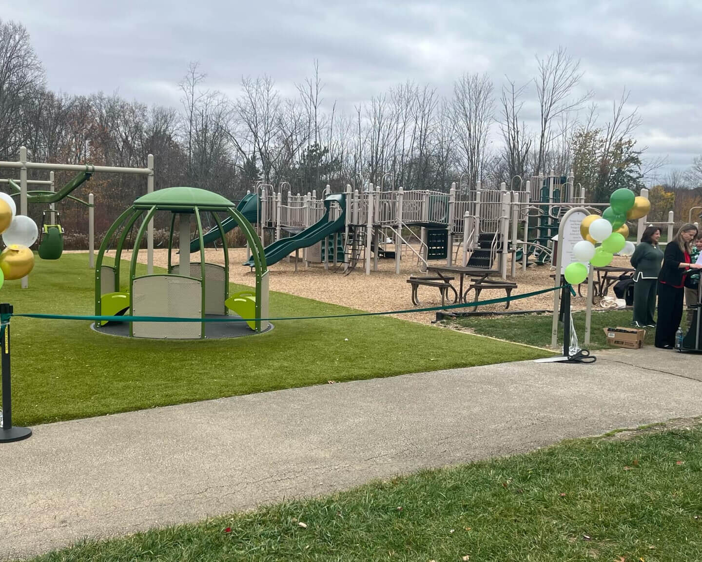 A playground with swings, slides, and a round green structure. Balloons decorate the area, and people stand near benches and tables on a cloudy day.