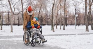 Two people in winter clothing, one in a wheelchair and the other standing behind, enjoy a snowy park path with leafless trees.